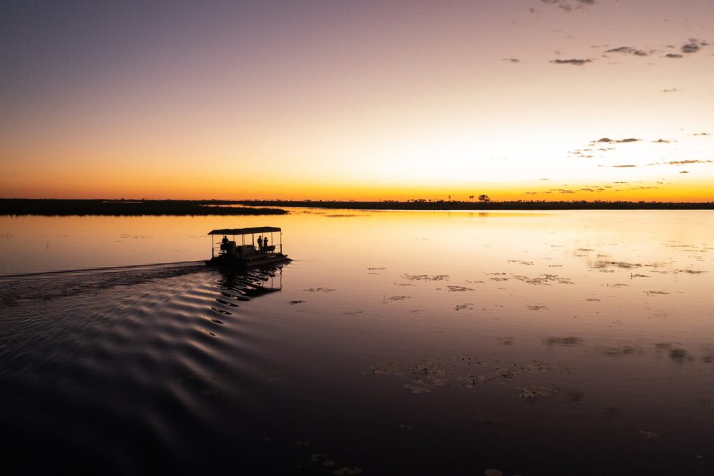 A pontoon boat with overhead canopy is silhouetted against a smooth lake, which mirrors the sunset in the clear sky above.