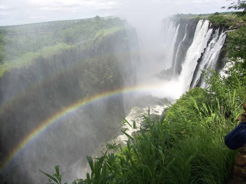 Victoria Falls cascades into a deep gorge framed by cliffs and lush vegetation, with a rainbow in the rising mist.