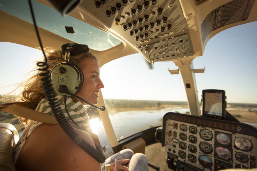 Woman wearing a headset smiles while piloting a helicopter, bathed in warm sunlight. The cockpit displays various controls, and a scenic view is visible.