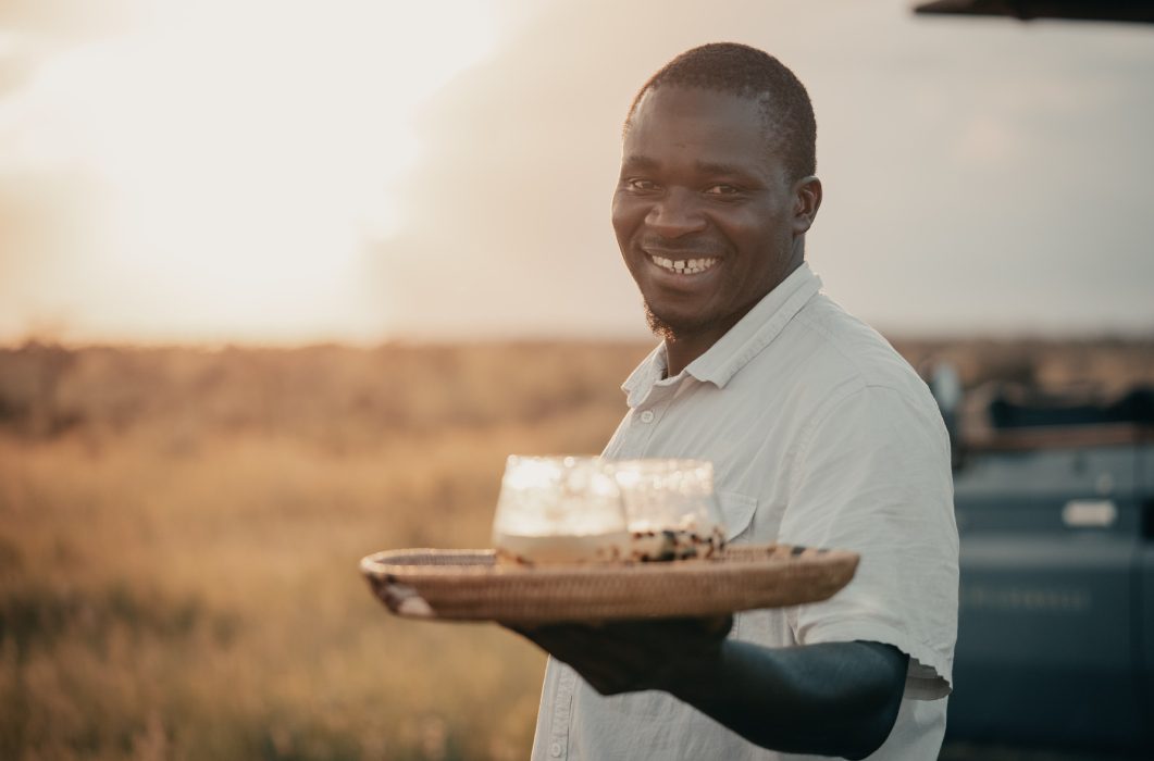 Smiling man holding a wooden tray with drinks in a sunlit field at a safari camp in Serengeti, Tanzania.