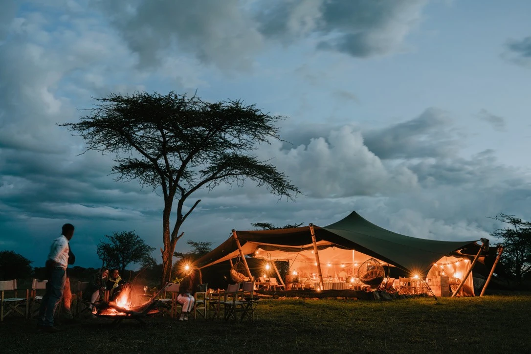 People gather around a campfire under a large acacia tree at dusk before a warmly lit tent, surrounded by a vast, cloudy sky.