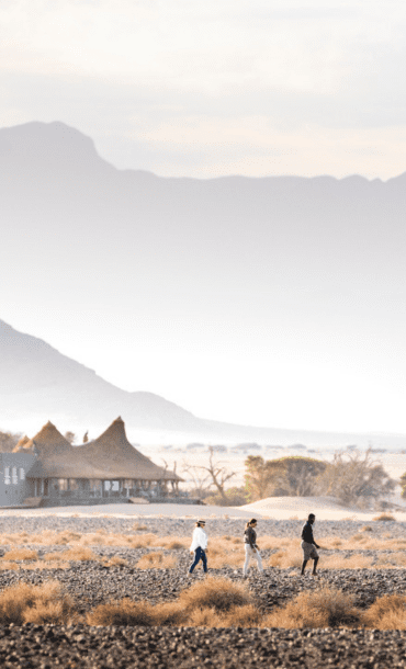 Three people traverse an arid landscape with safari camp in the distance with a dramatic peaked roof, and towering moutains in the distance.