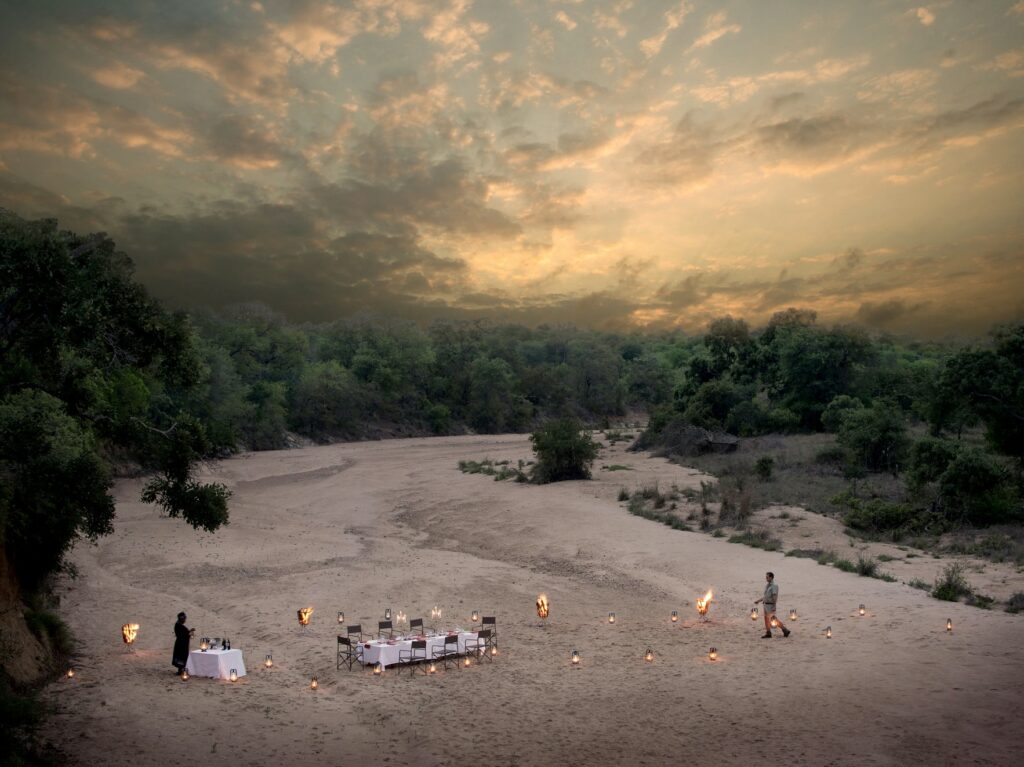 Two waiters attend to a fine dining table set up for a bush dinner in a dry, sandy riverbed, with tiki torches, at sunset.