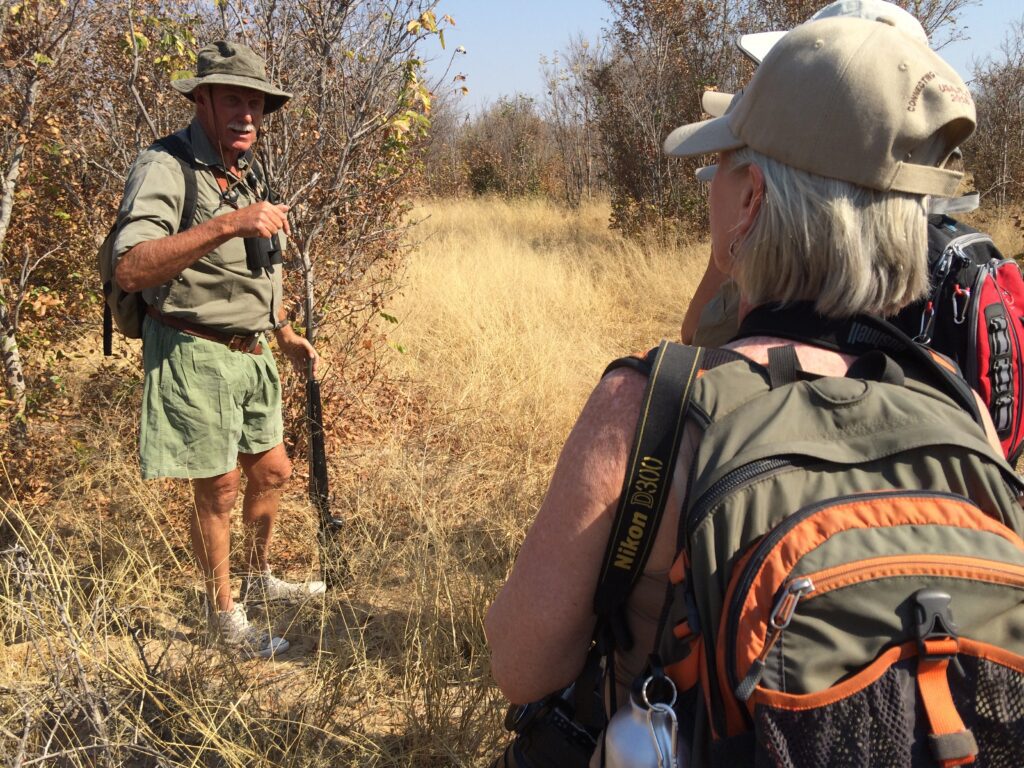 A safari guide wearing a bucket hat and carrying a rifle, speaking to a group of travelers on a bush walk.