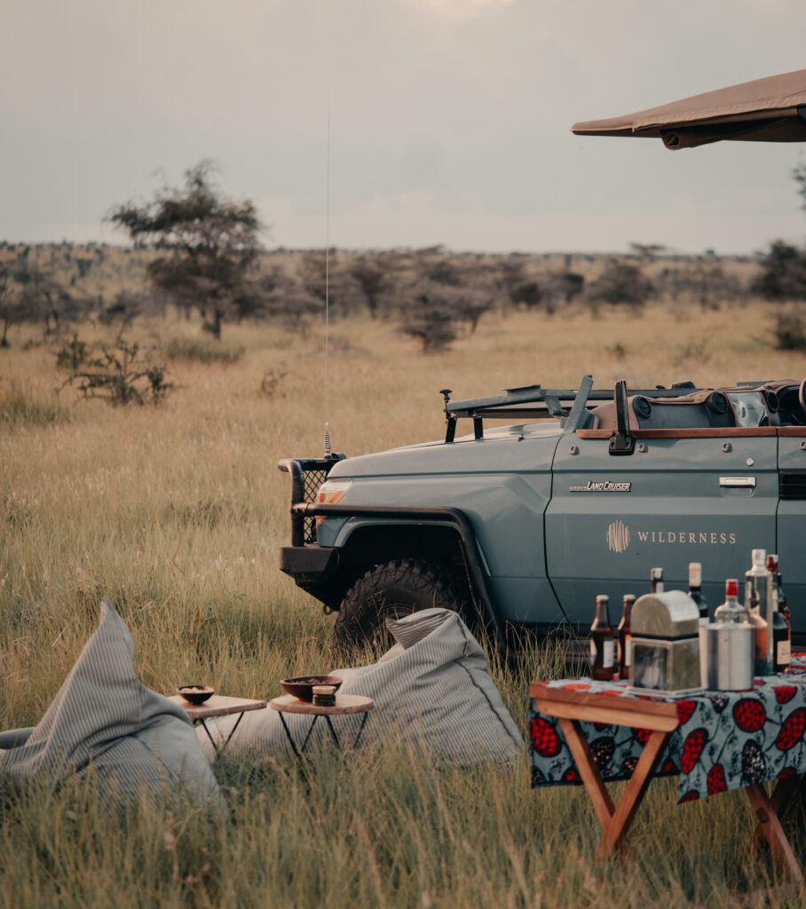 A safari vehicle is parked next to a sundowner setup, with bar items on a folding teak table, and beanbag chairs for lounging.