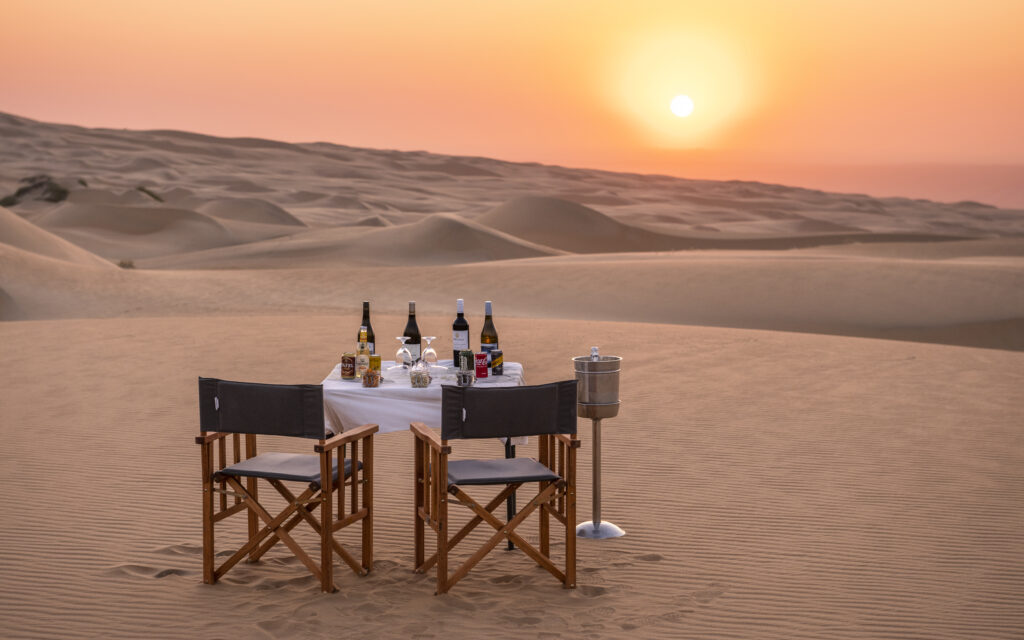Camp chairs and a small table with a white tablecloth is set with bottles of wine and drinks, in a desert dune landscape, at sunset.