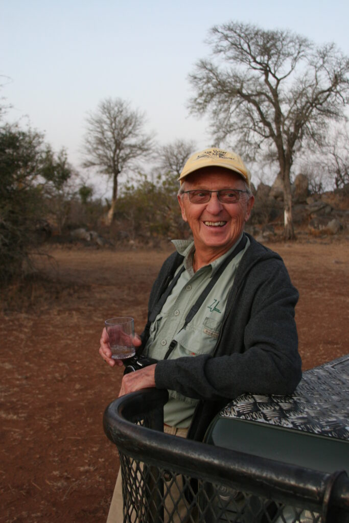 An smiling elderly man holds a drink while leaning on a safari vehicle. 