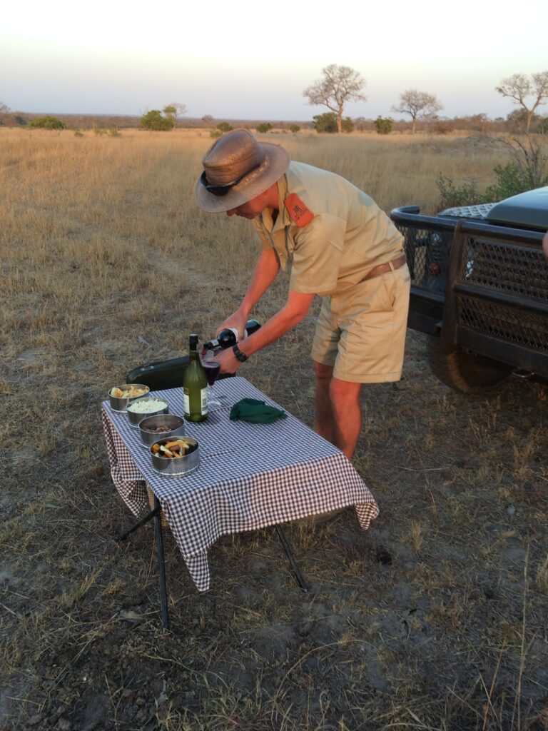 A safari guide in traditional guiding attire prepares drinks on a camp table set up in front of his Land Cruiser.