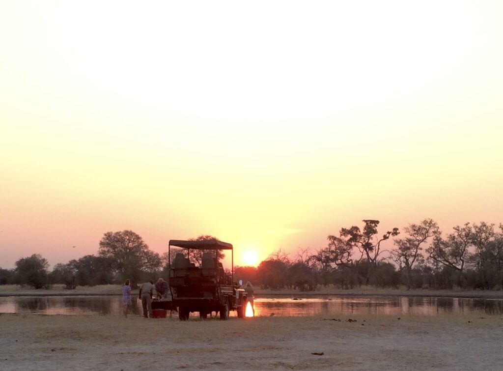 Safari vehicle parked in front of a small pond at sunset with a few people standing around the front, enjoying drinks and socializing.