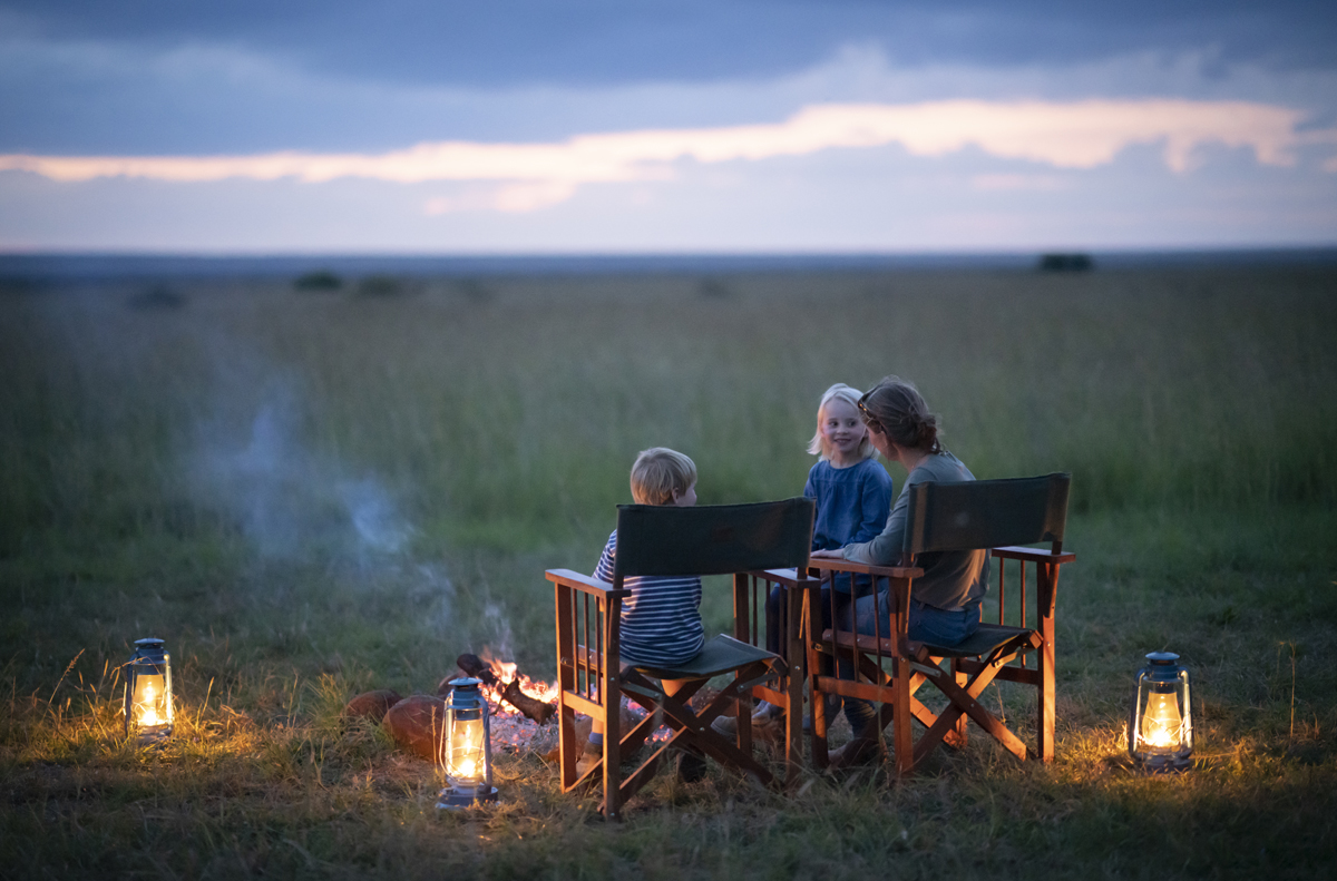A mother and two children sit in camp chairs in front of a campfire, surrounded by lanterns, as the sun sets.