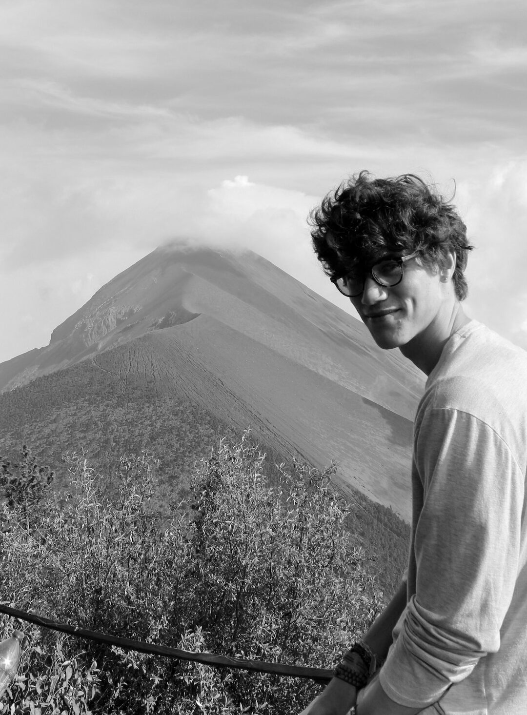 Black and white image of a young man smiling at the camera with a dramatic peak in the background.