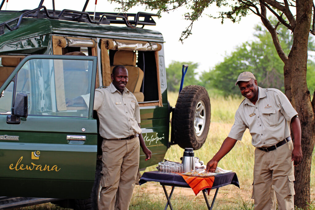 Two safari guides smile while setting up a small table next to a safari vehicle with drinks and snacks.