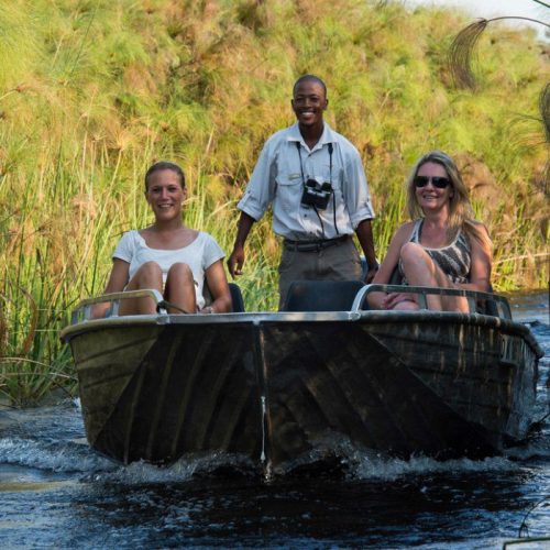Three people in a small boat navigate through lush greenery on a river. Two women sit smiling in front, while a man at the back steers, looking content.