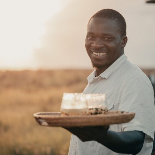 Smiling man holding a wooden tray with drinks in a sunlit field at a safari camp in Serengeti, Tanzania.