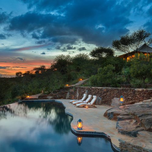 An infinity pool at a safari camp in Loisaba, Kenya surrounded by natural stone with a colorful sunset in the distance.