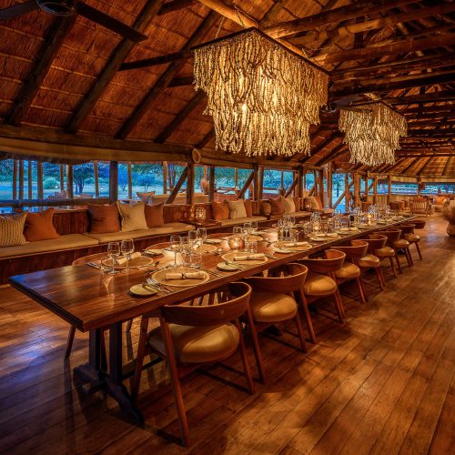 A long wooden dining table is set for dinner, in a rustic thatch lodge, with dramatic crystal chandeliers overhead, creating a scene both elegant and rustic.