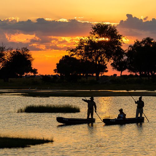 Two Botswana mekoro being poled through a large lagoon are silouetted against a dramatic sunset.