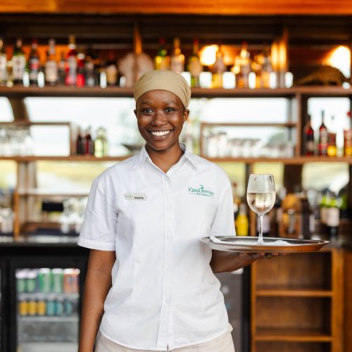 A smiling woman dressed in a white uniform holds a serving tray in front of a rusting wooden bar.