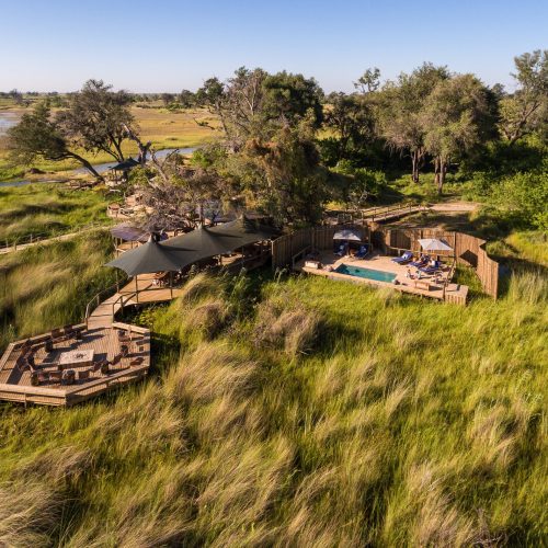 Aerial scene of a luxury safari camp with plunge pool and observation decks, overlooking a grassy plain and wetlands.