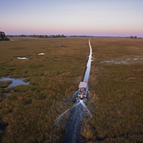 A canopied motor boat navigates a narrow channel in a grassy wetland, under a purple sunset sky, evoking a remote feeling.