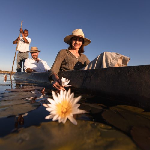 Two passengers seated in a traditional Botswanan mokoro, a guide poling in the back, and blooming lily pads under a clear blue sky.