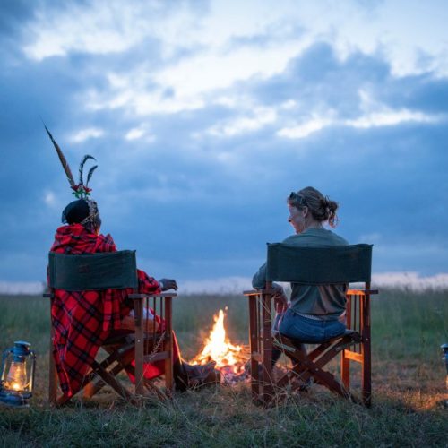 A female tourist and a Samburu warrior in traditional dress talking near a campfire, surrounded by lanterns, at dusk on a Kenyan savanna.