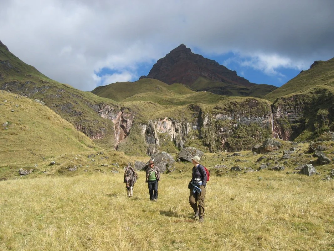 Two hikers and a donkey cross a grassy valley with rocky cliffs under a partly cloudy sky, a towering peak in the background.